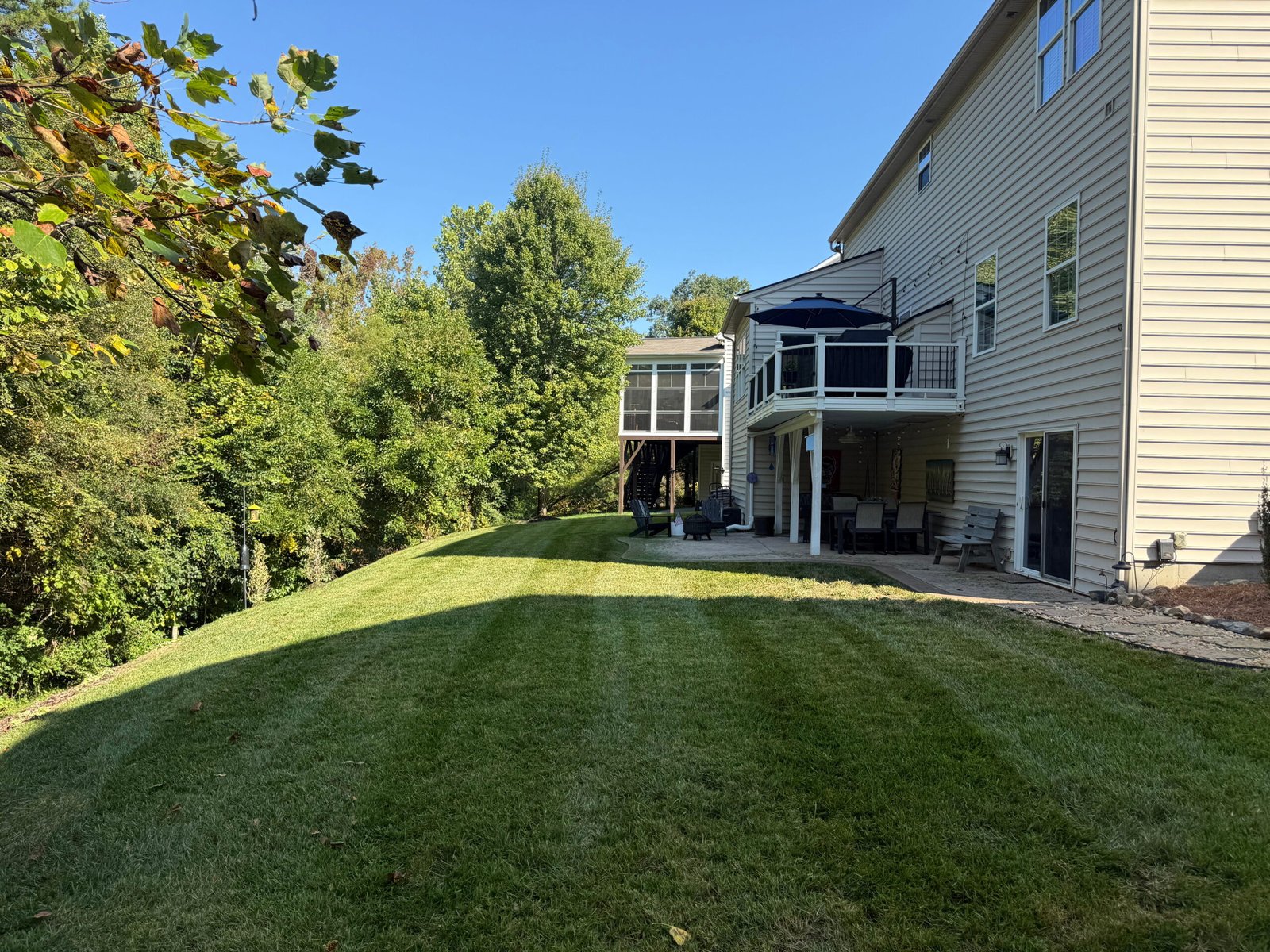 Expertly mowed sloped backyard with crisp lawn stripes bordering a wooded area and a residential patio deck.